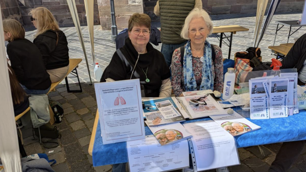 Zwei Frauen der Regionalgruppe Franken informieren mit einem Marktstand in Nürnberg über die Lungenfibrose. Auf dem Tisch vor ihnen iegen Flyer und Broschüren aus.