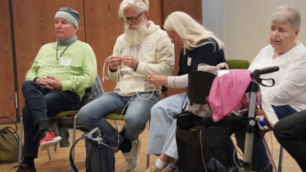 Mobiles Sauerstoffgerät Eine Veranstaltung des Lungenfibrose e.V. Fünf Personen sitzen in einem Veranstaltungsraum. Eine Teilnehmerin mit Sauerstoffgerät und Rollator ist dabei. Zwei Teilnehmende machen sich mit einem mobilen Sauerstoffgerät mit Schlauch vertraut. Das Gerät befindet sich in einem kleinen Rucksack.