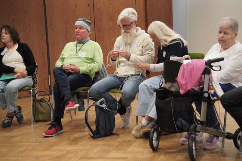 Mobiles Sauerstoffgerät Eine Veranstaltung des Lungenfibrose e.V. Fünf Personen sitzen in einem Veranstaltungsraum. Eine Teilnehmerin mit Sauerstoffgerät und Rollator ist dabei. Zwei Teilnehmende machen sich mit einem mobilen Sauerstoffgerät mit Schlauch vertraut. Das Gerät befindet sich in einem kleinen Rucksack.